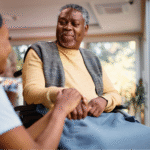 A young Black female caregiver holding the hand of a Black elderly man in a wheelchair, illustrating the compassion involved in caring for a loved one with Alzheimer's