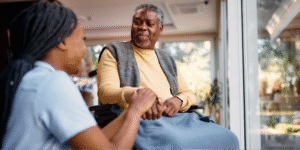 A young Black female caregiver holding the hand of a Black elderly man in a wheelchair, illustrating the compassion involved in caring for a loved one with Alzheimer's