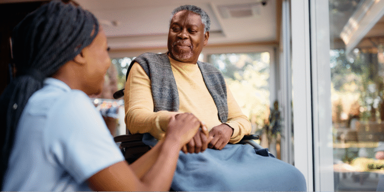 A young Black female caregiver holding the hand of a Black elderly man in a wheelchair, illustrating the compassion involved in caring for a loved one with Alzheimer's