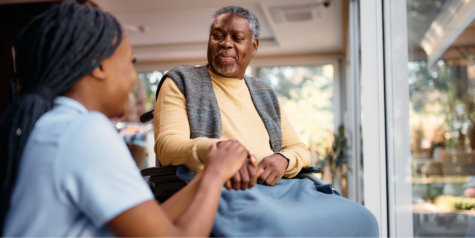A young Black female caregiver holding the hand of a Black elderly man in a wheelchair, illustrating the compassion involved in caring for a loved one with Alzheimer's
