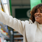 A Black woman with curly hair smiling while selecting yogurt from a refrigerated supermarket shelf of probiotic foods.