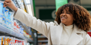 A Black woman with curly hair smiling while selecting yogurt from a refrigerated supermarket shelf of probiotic foods.