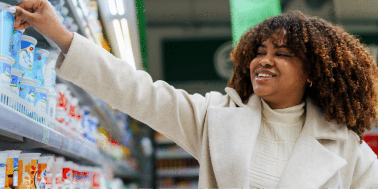 A Black woman with curly hair smiling while selecting yogurt from a refrigerated supermarket shelf of probiotic foods.