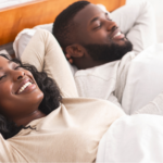 A young Black couple lying in bed with their eyes closed and smiles on their faces, preparing for better sleep.
