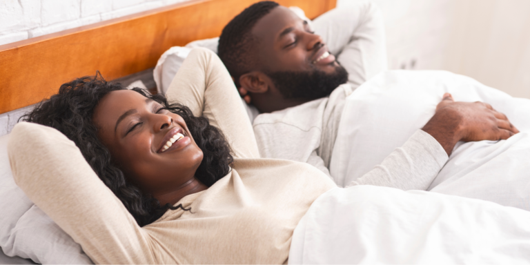 A young Black couple lying in bed with their eyes closed and smiles on their faces, preparing for better sleep.