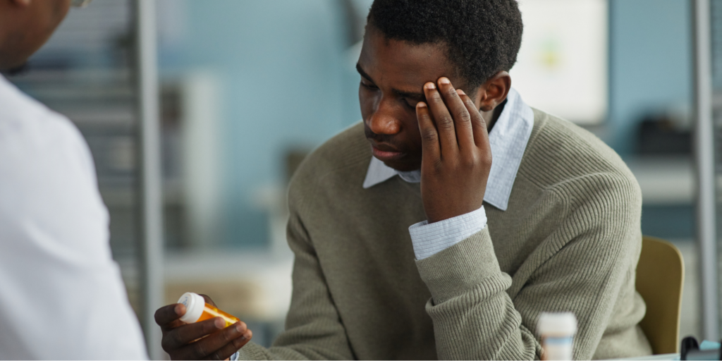 Young adult Black man sitting in a medical office holding a prescription bottle while massaging forehead with his other hand, and consulting with the doctor about his high blood pressure medication.