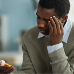 Young adult Black man sitting in a medical office holding a prescription bottle while massaging forehead with his other hand, and consulting with the doctor about his high blood pressure medication.