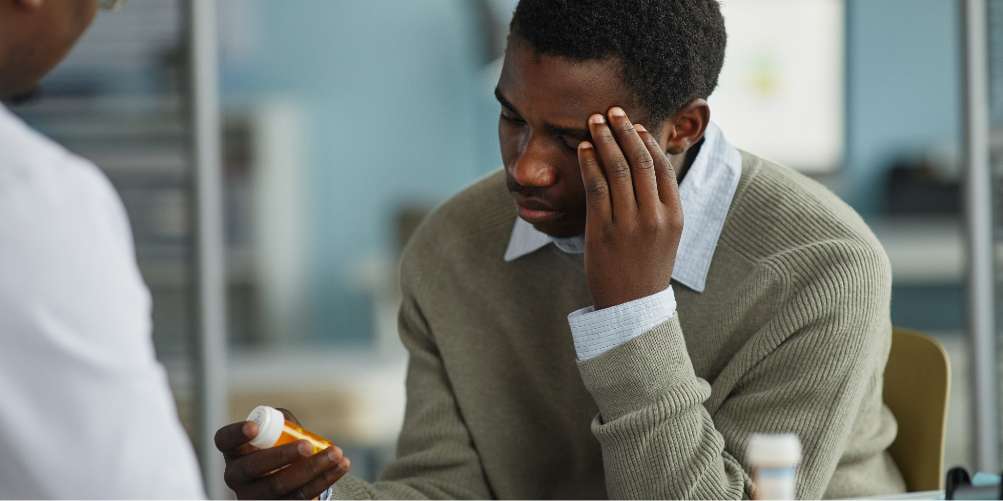 Young adult Black man sitting in a medical office holding a prescription bottle while massaging forehead with his other hand, and consulting with the doctor about his high blood pressure medication.