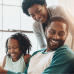 Happy Black family, including father, mother, daughter, and son, sitting on a white couch reading a book on autism.