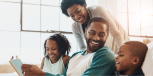 Happy Black family, including father, mother, daughter, and son, sitting on a white couch reading a book on autism.