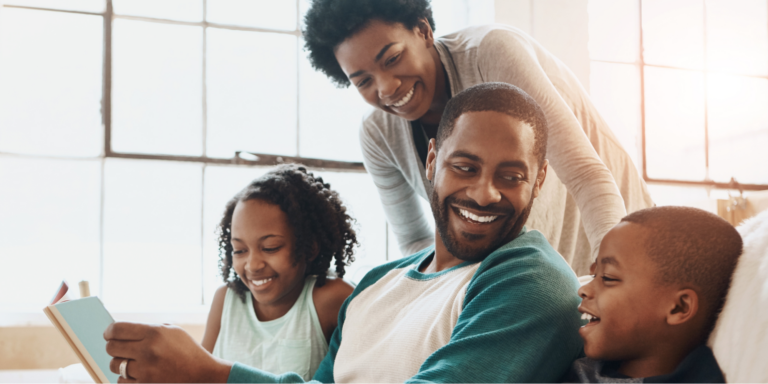 Happy Black family, including father, mother, daughter, and son, sitting on a white couch reading a book on autism.