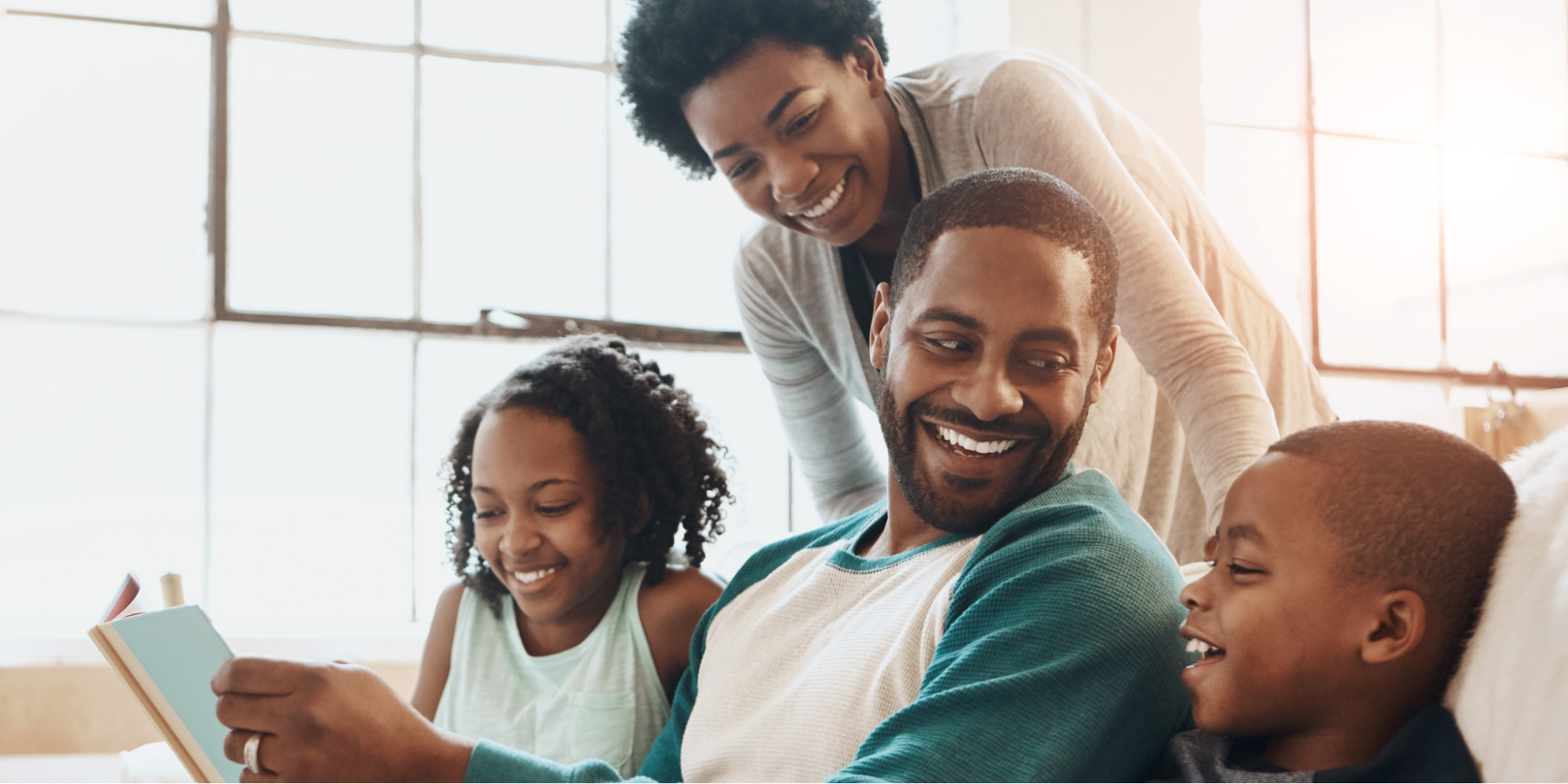 Happy Black family, including father, mother, daughter, and son, sitting on a white couch reading a book on autism.