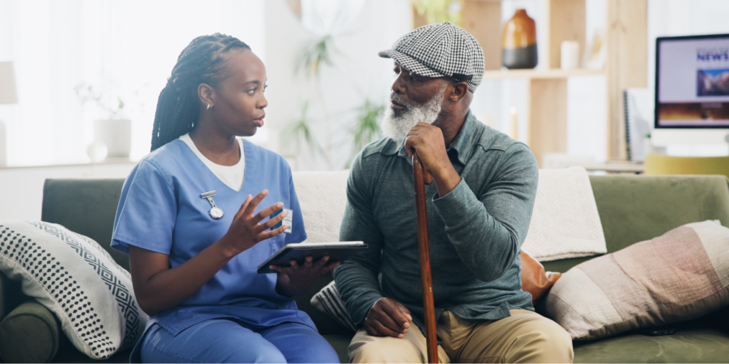 Black elderly man sitting on the couch, holding a cane, and learning about the Medicare Annual Wellness Visit and cognitive assessments with his nurse.
