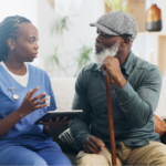 Black elderly man sitting on the couch, holding a cane, and learning about the Medicare Annual Wellness Visit and cognitive assessments with his nurse.
