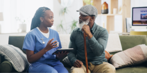 Black elderly man sitting on the couch, holding a cane, and learning about the Medicare Annual Wellness Visit and cognitive assessments with his nurse.