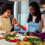 A smiling Black woman in denim overalls teaching her children how to chop vegetables like broccoli and carrots to prep quick anti-inflammatory meals.