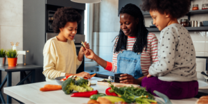 A smiling Black woman in denim overalls teaching her children how to chop vegetables like broccoli and carrots to prep quick anti-inflammatory meals.