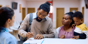 A Black family standing at a clinic reception desk, where the father is filling out medical paperwork for prostate cancer treatment.