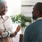 A smiling Black healthcare professional in a headwrap holding a stethoscope while talking to a male patient about heart failure medication.