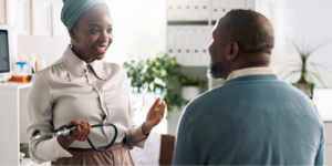 A smiling Black healthcare professional in a headwrap holding a stethoscope while talking to a male patient about heart failure medication.