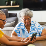 Two senior African American women in glasses, sitting at a table, reading an article about immunization shots on their smartphone.