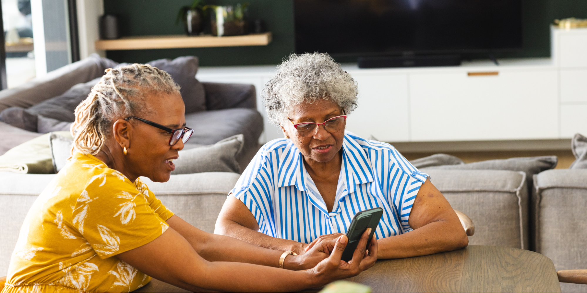 Two senior African American women in glasses, sitting at a table, reading an article about immunization shots on their smartphone.