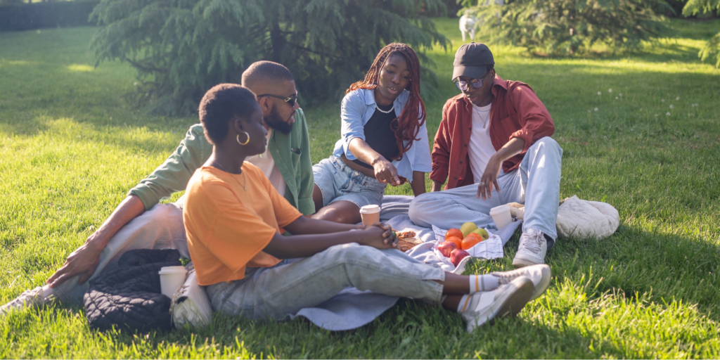 A group of four young Black friends enjoying a picnic with fresh fruit and vegetables for their gut health on a sunny park lawn.