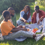 A group of four young Black friends enjoying a picnic with fresh fruit and vegetables for their gut health on a sunny park lawn.