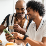 Two young Black men in collared shirts drinking coffee and eating breakfast while reading an article about hidden sugars and fat on a cell phone.