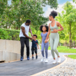 A Black family in athletic wear exercises in a park. The father wears a white tank top and black pants, the young son has a navy t-shirt, the daughter wears a lavender-blue unitard, and the mother wears a sage green sports bra and leggings set, illustrating what to look for every day to keep your partner’s heart strong.