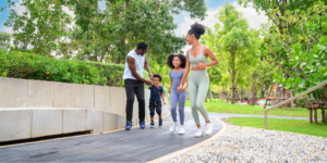 A Black family in athletic wear exercises in a park. The father wears a white tank top and black pants, the young son has a navy t-shirt, the daughter wears a lavender-blue unitard, and the mother wears a sage green sports bra and leggings set, illustrating what to look for every day to keep your partner&rsquo;s heart strong.