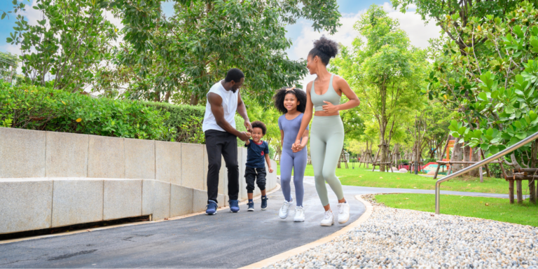 A Black family in athletic wear exercises in a park. The father wears a white tank top and black pants, the young son has a navy t-shirt, the daughter wears a lavender-blue unitard, and the mother wears a sage green sports bra and leggings set, illustrating what to look for every day to keep your partner’s heart strong.