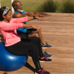 An older Black couple exercising on a sunny outdoor wooden deck next to a pool. The woman is seated on a large blue stability ball wearing a pink long-sleeved shirt and black leggings, while the man sits next to her in a blue tank top and black shorts. Both have their arms extended forward, performing focused at-home exercises to improve after surgery bladder control.