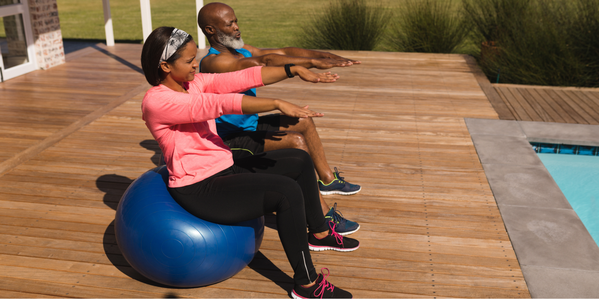 An older Black couple exercising on a sunny outdoor wooden deck next to a pool. The woman is seated on a large blue stability ball wearing a pink long-sleeved shirt and black leggings, while the man sits next to her in a blue tank top and black shorts. Both have their arms extended forward, performing focused at-home exercises to improve after surgery bladder control.