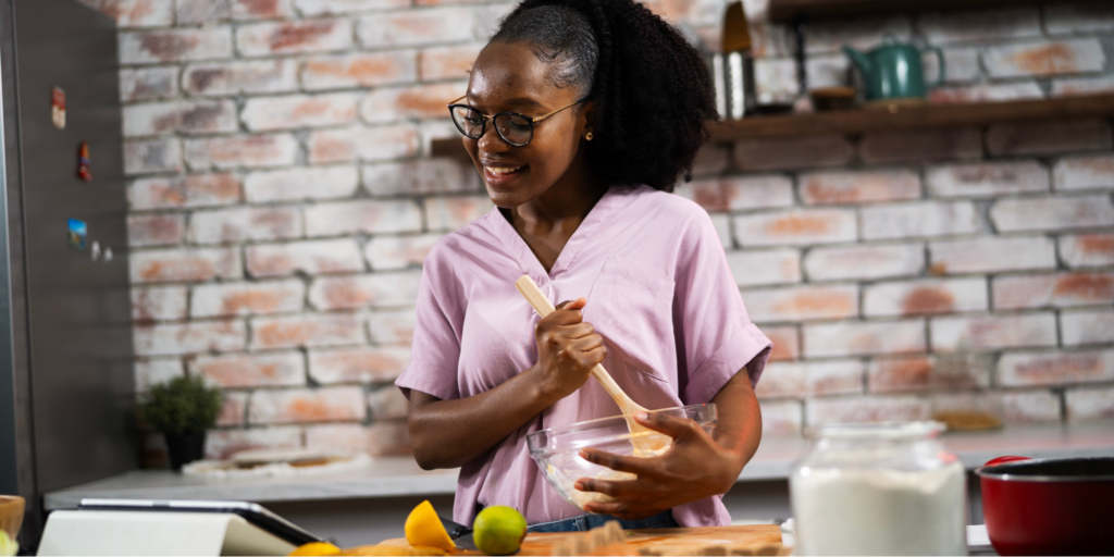 A young Black woman with glasses and her hair in a ponytail wears a light purple V-neck shirt while smiling and mixing ingredients in a glass bowl with a wooden spoon. She is following a gut health recipe on a table, as part of her morning gut routine, in a rustic kitchen with a brick backsplash.