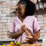 A young Black woman with glasses and her hair in a ponytail wears a light purple V-neck shirt while smiling and mixing ingredients in a glass bowl with a wooden spoon. She is following a gut health recipe on a table, as part of her morning gut routine, in a rustic kitchen with a brick backsplash.