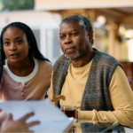 An older Black man with gray hair and a beard, wearing a yellow sweater and a patterned vest, sits at an outdoor table holding a wooden cane. Next to him, a younger Black woman with long braids and a pink shirt leans in with a concerned expression as they listen to a man in a blue shirt review medical information about blood pressure and stroke.