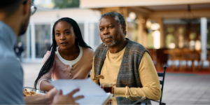 An older Black man with gray hair and a beard, wearing a yellow sweater and a patterned vest, sits at an outdoor table holding a wooden cane. Next to him, a younger Black woman with long braids and a pink shirt leans in with a concerned expression as they listen to a man in a blue shirt review medical information about blood pressure and stroke.