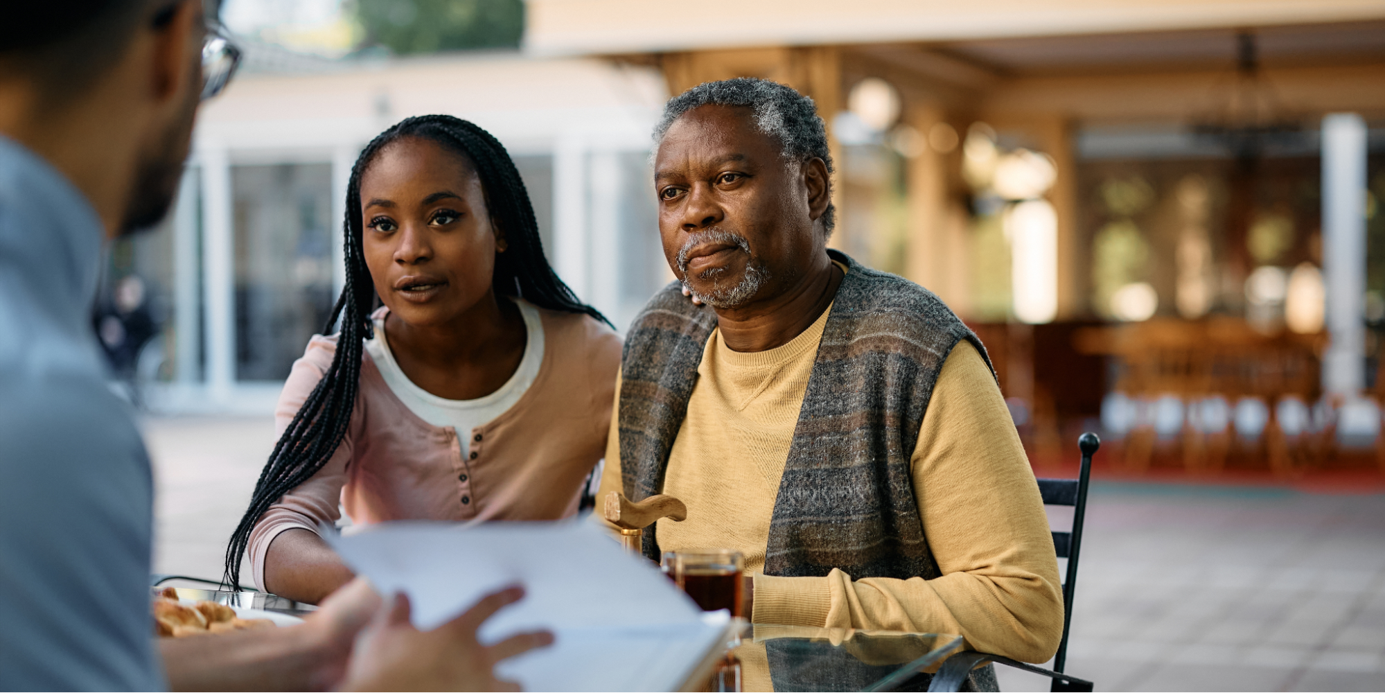 An older Black man with gray hair and a beard, wearing a yellow sweater and a patterned vest, sits at an outdoor table holding a wooden cane. Next to him, a younger Black woman with long braids and a pink shirt leans in with a concerned expression as they listen to a man in a blue shirt review medical information about blood pressure and stroke.