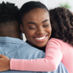 A Black woman with breast cancer with a serene smile and eyes closed, is hugged by her partner, seen from the back in a blue denim shirt, and her young daughter with curly hair, wearing a pink long-sleeved shirt, demonstrating breast cancer community support.