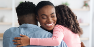 A Black woman with breast cancer with a serene smile and eyes closed, is hugged by her partner, seen from the back in a blue denim shirt, and her young daughter with curly hair, wearing a pink long-sleeved shirt, demonstrating breast cancer community support.