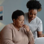 An older Black woman with short hair and a brown V-neck sweater sits at a table, looking at a laptop screen with her hand near her chin. Standing next to her, a Black female healthcare worker in light gray scrubs leans in to point at the screen to review insurance paperwork, quit-smoking medication options, and a phone with a quitline number, representing how to use insurance benefits to quit smoking.