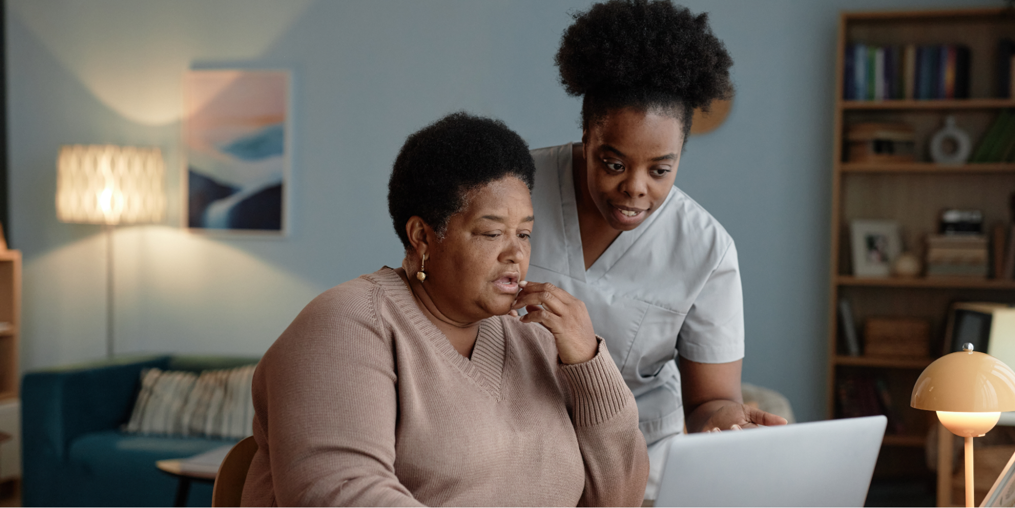 An older Black woman with short hair and a brown V-neck sweater sits at a table, looking at a laptop screen with her hand near her chin. Standing next to her, a Black female healthcare worker in light gray scrubs leans in to point at the screen to review insurance paperwork, quit-smoking medication options, and a phone with a quitline number, representing how to use insurance benefits to quit smoking.