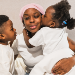 A Black woman with lupus wearing a pink headwrap and a white t-shirt sits in bed, sharing a tender moment with her two young children. Her daughter, in a white shirt with blue hair clips, kisses her cheek while her son leans in close for a hug.