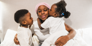 A Black woman with lupus wearing a pink headwrap and a white t-shirt sits in bed, sharing a tender moment with her two young children. Her daughter, in a white shirt with blue hair clips, kisses her cheek while her son leans in close for a hug.