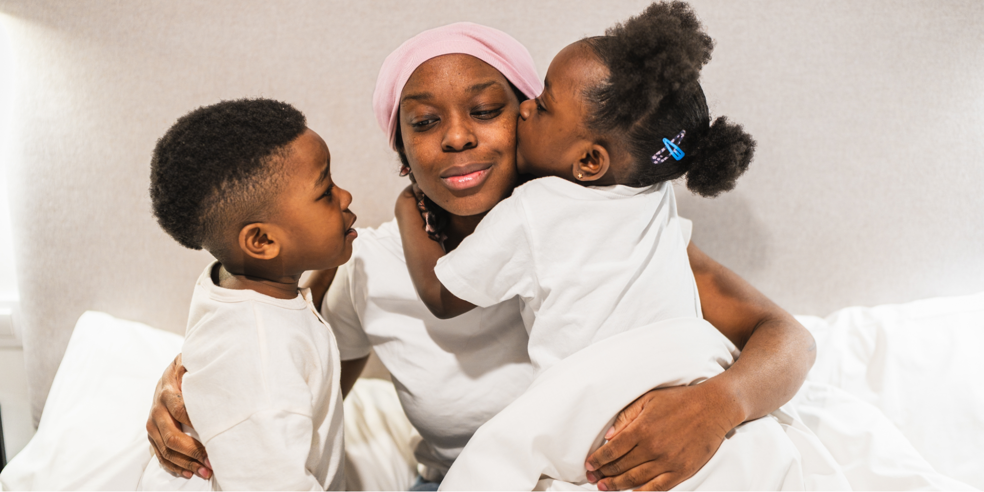 A Black woman with lupus wearing a pink headwrap and a white t-shirt sits in bed, sharing a tender moment with her two young children. Her daughter, in a white shirt with blue hair clips, kisses her cheek while her son leans in close for a hug.