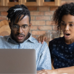 A young Black couple reading an article about hantavirus on their laptop screen with expressions of shock and alarm in their kitchen. The man, wearing glasses and a light patterned shirt, has his mouth wide open, while the woman, in a dark floral top, gasps with her hand on his shoulder.