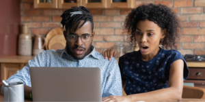 A young Black couple reading an article about hantavirus on their laptop screen with expressions of shock and alarm in their kitchen. The man, wearing glasses and a light patterned shirt, has his mouth wide open, while the woman, in a dark floral top, gasps with her hand on his shoulder.