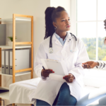 A Black female doctor with her hair in a high ponytail, wearing a white lab coat and stethoscope, listens intently while holding a clipboard. She is consulting with a Black woman in a striped long-sleeve shirt and jeans, who is gesturing toward her stomach with a concerned expression asking the doctor questions before starting a new UC treatment.