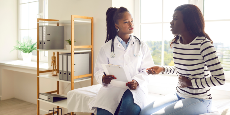Can Gut Issues Cause Cancer? 2 A Black female doctor with her hair in a high ponytail, wearing a white lab coat and stethoscope, listens intently while holding a clipboard. She is consulting with a Black woman in a striped long-sleeve shirt and jeans, who is gesturing toward her stomach with a concerned expression asking the doctor questions before starting a new UC treatment.